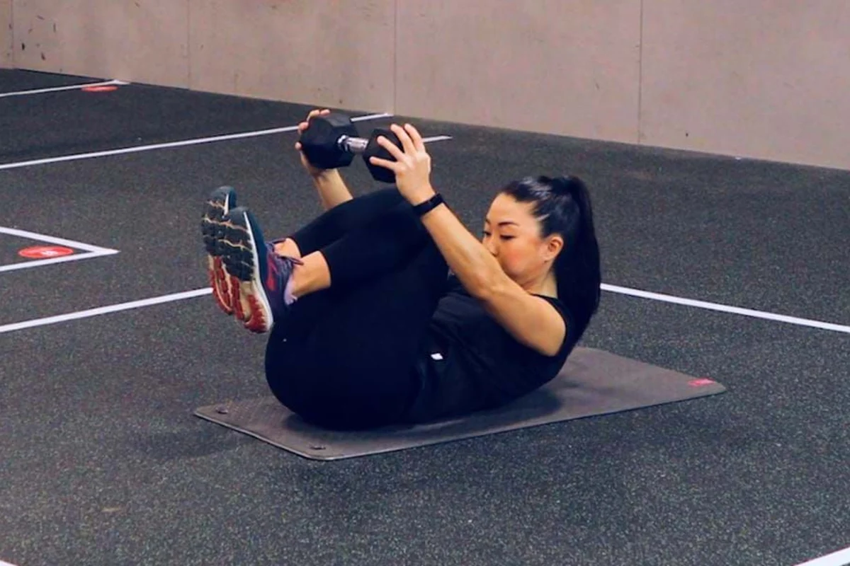 A beginner lying on a mat holding a hollow body tuck, knees drawn in and shoulders slightly lifted while the ribs stay down and the low back stays connected to the mat