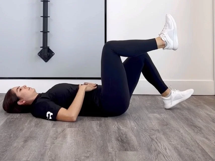 A beginner lying on a mat performing heel taps, alternating feet to lightly touch the floor while keeping the ribs down and the shins mostly parallel to the floor