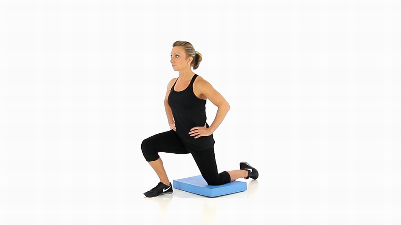 A person doing a half-kneeling hip flexor stretch on a yoga mat at home, one knee on a folded towel, torso tall, hands resting on the front thigh