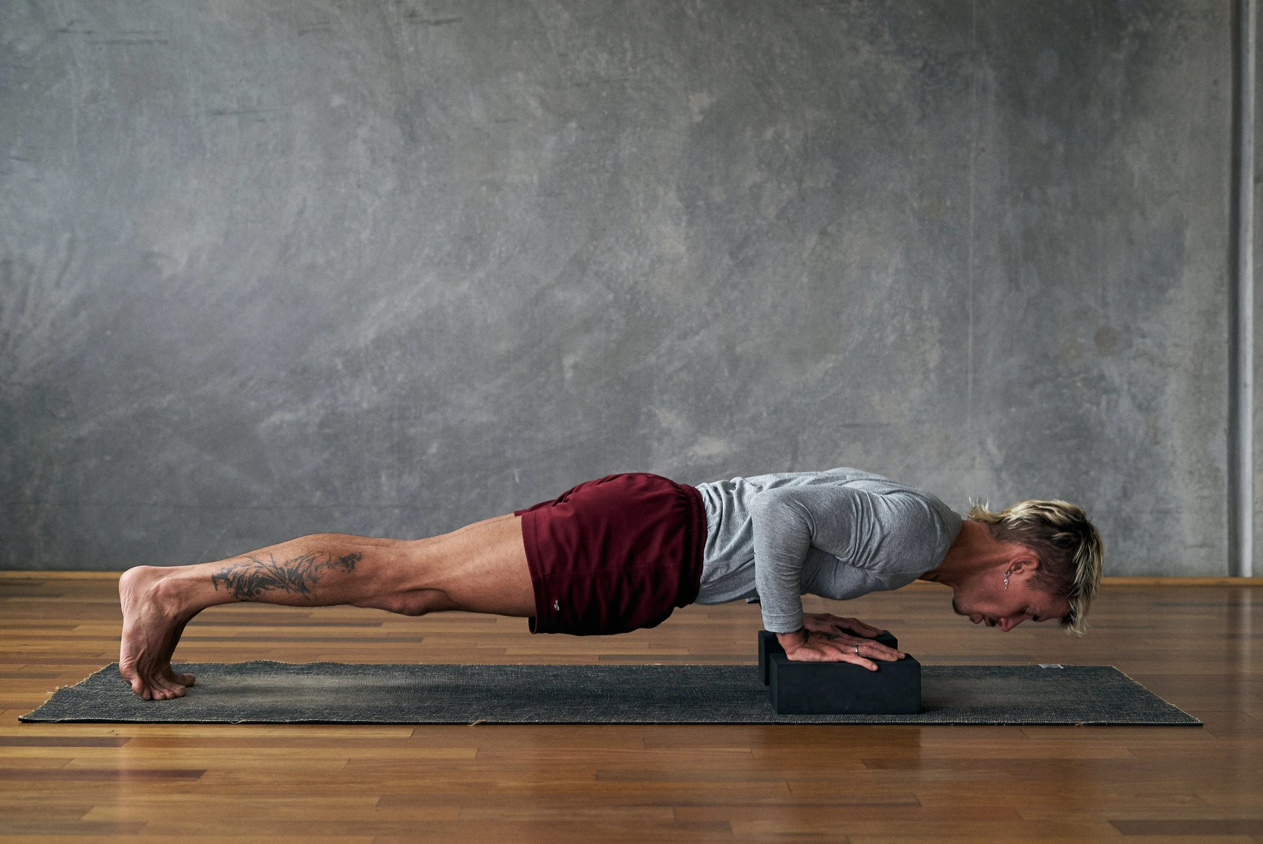 A person doing an assisted one-arm push-up with one hand on the floor and the other supported on a yoga block, feet wide for stability