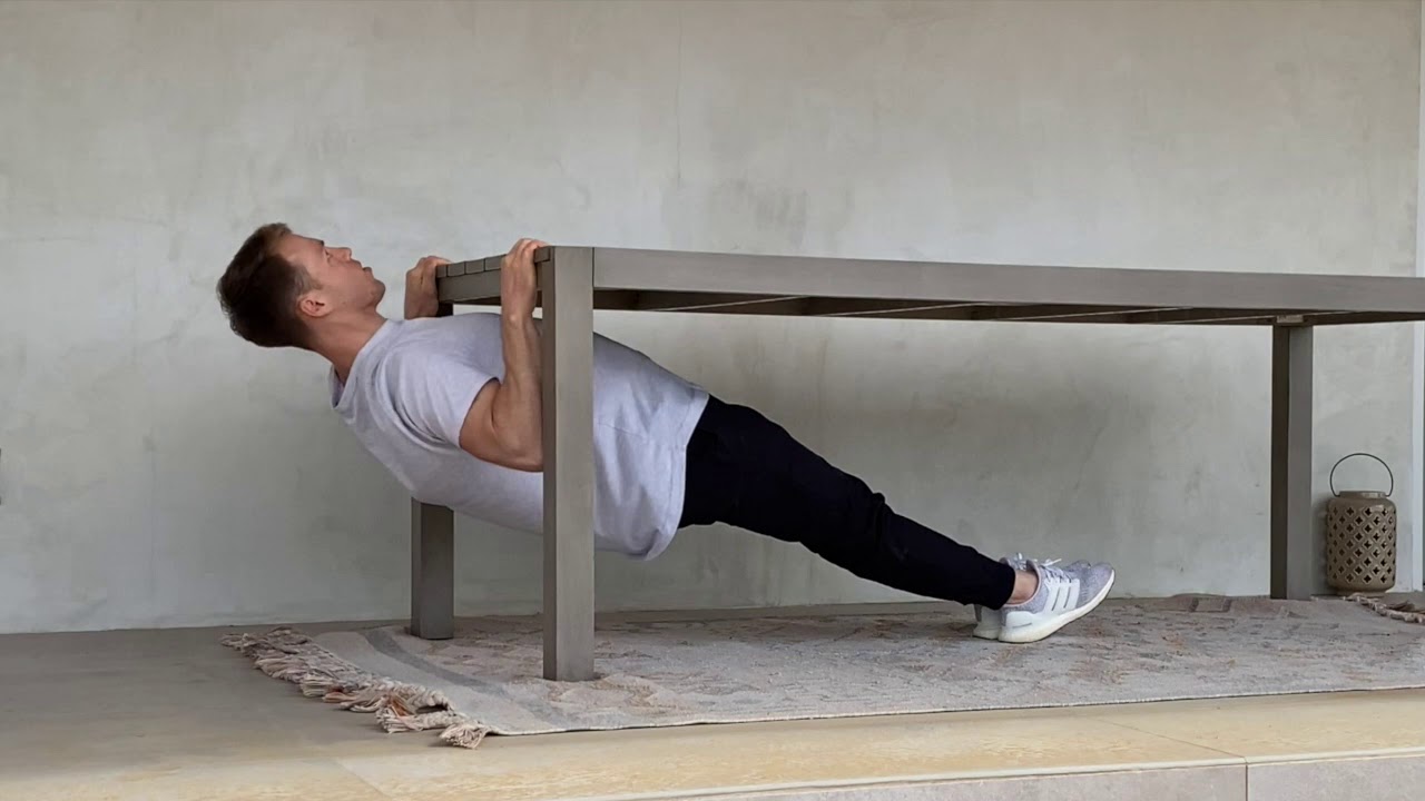 A person doing an inverted row under a sturdy dining table, gripping the table edge with knees bent and heels on the floor