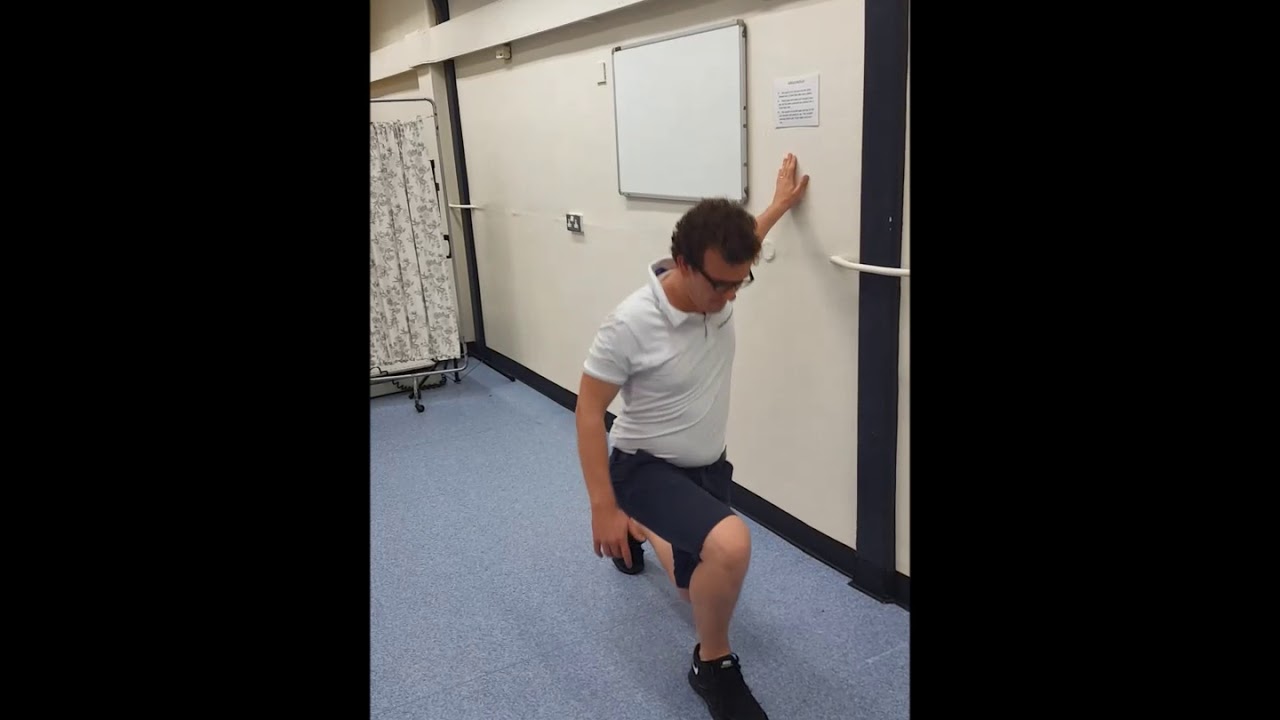 A person performing a reverse lunge while lightly holding a wall for balance in a hallway
