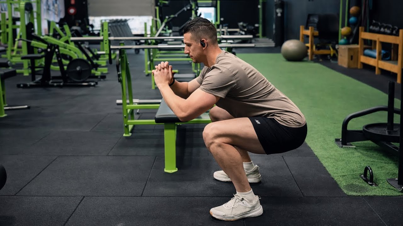 A person performing a slow bodyweight squat at home, pausing near the bottom with arms extended for balance