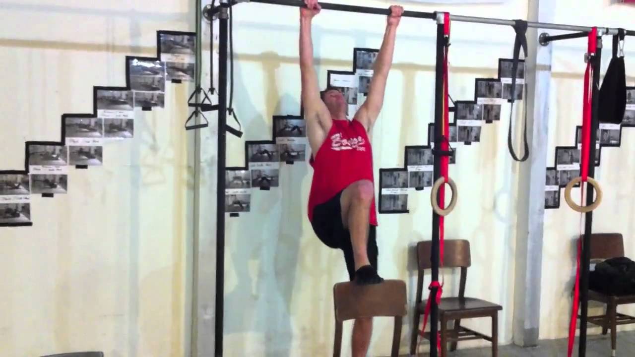 A person standing on a chair under a doorway pull-up bar, starting at the top position and lowering slowly into a negative pull-up