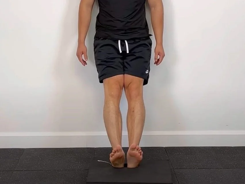 A person standing upright near a wall, heels grounded while the front of both feet lifts so the toes point upward during a toe raise